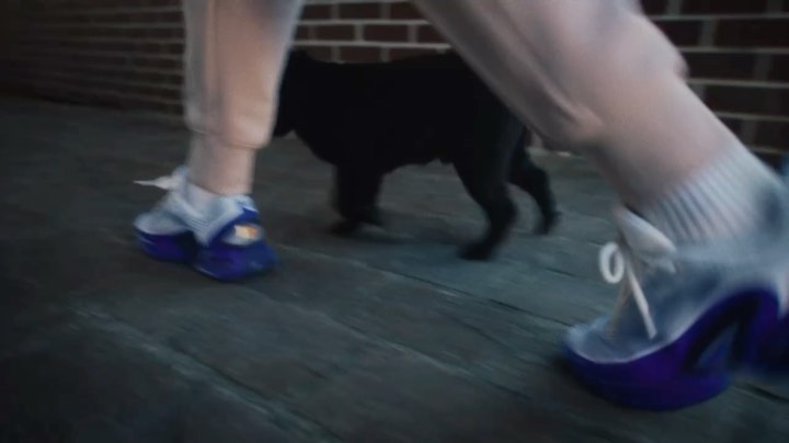 Person wearing white and purple sneakers walking next to a black cat on a wooden porch outside a brick building.