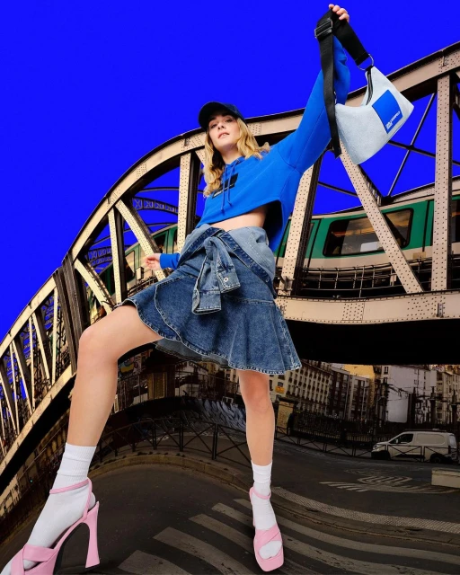 Stylish young woman in denim skirt, blue hoodie, and pink heels poses with a blue bag on a bridge with a train passing in the background.
