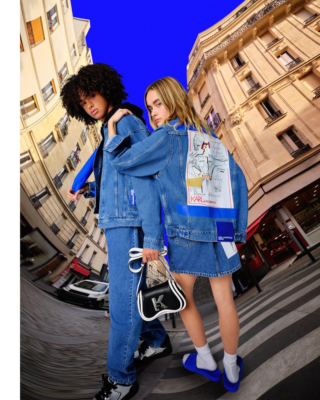 Two fashion models in denim outfits pose on a city street with Parisian buildings. One carries a black purse and wears blue sandals.