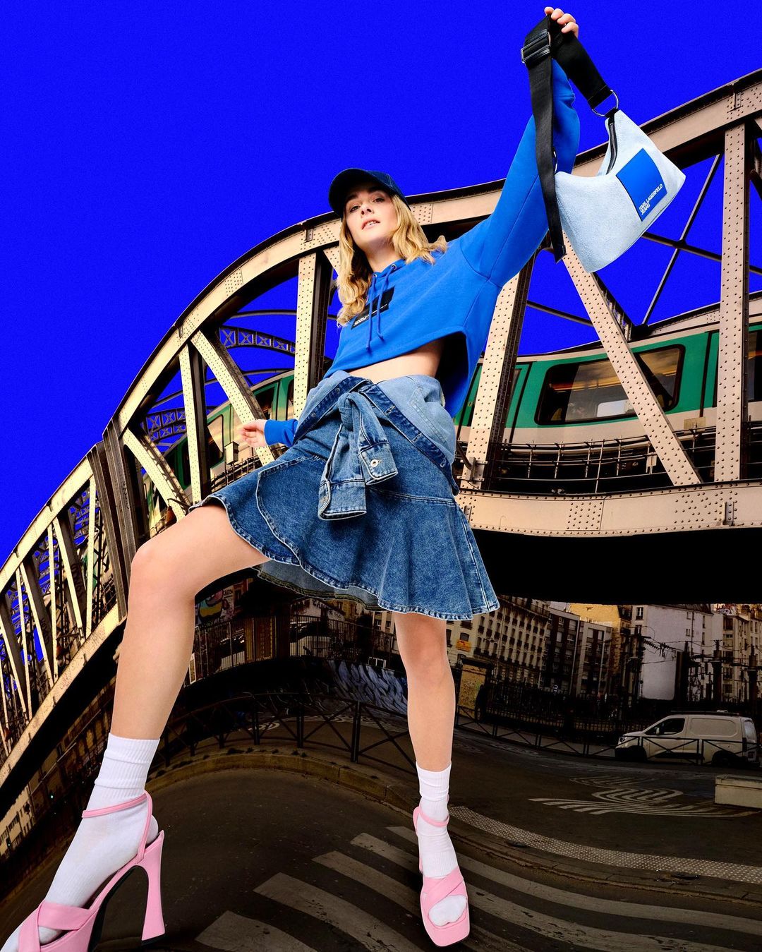 A woman poses on a city street with an elevated train in the background wearing a cropped blue sweatshirt and a denim skirt.