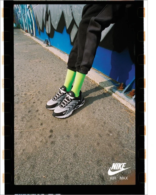 A person wearing black sweatpants and bright green socks is sitting near graffiti art with Nike Air Max shoes visible in the foreground.