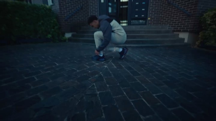 Young man kneels on a cobblestone path, tying his shoelaces in front of brick building steps, a lush bush, and dim, overcast lighting.