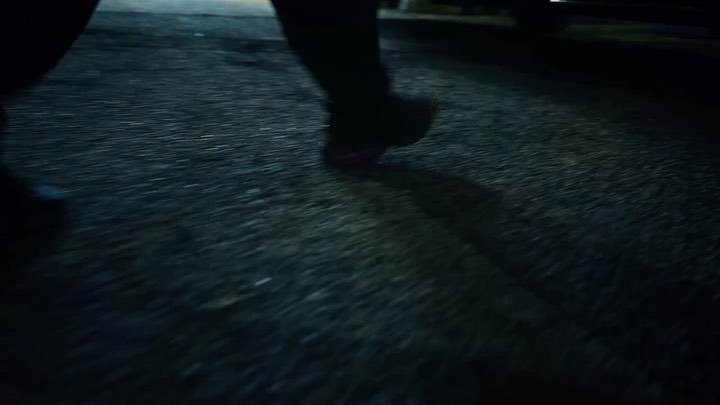 Close-up of a person walking on an asphalt street at night, with their shadow visible and a car parked in the distance.