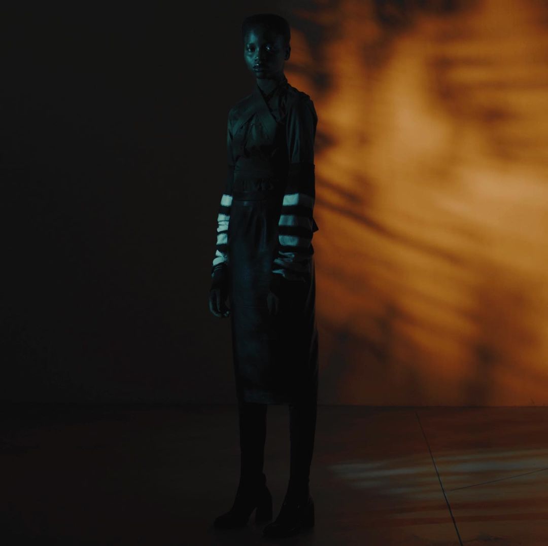A full-length portrait of a Black woman in a dark outfit, under moody lighting with an orange background, showcases a unique studio shot.