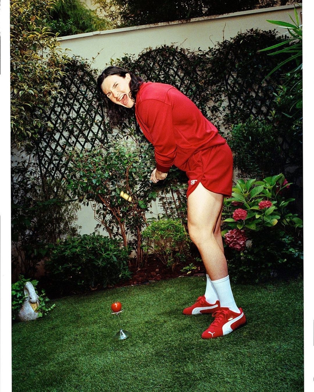 Adult female in red athletic wear with white socks and Puma sneakers plays croquet on the green grass in a lush outdoor setting.