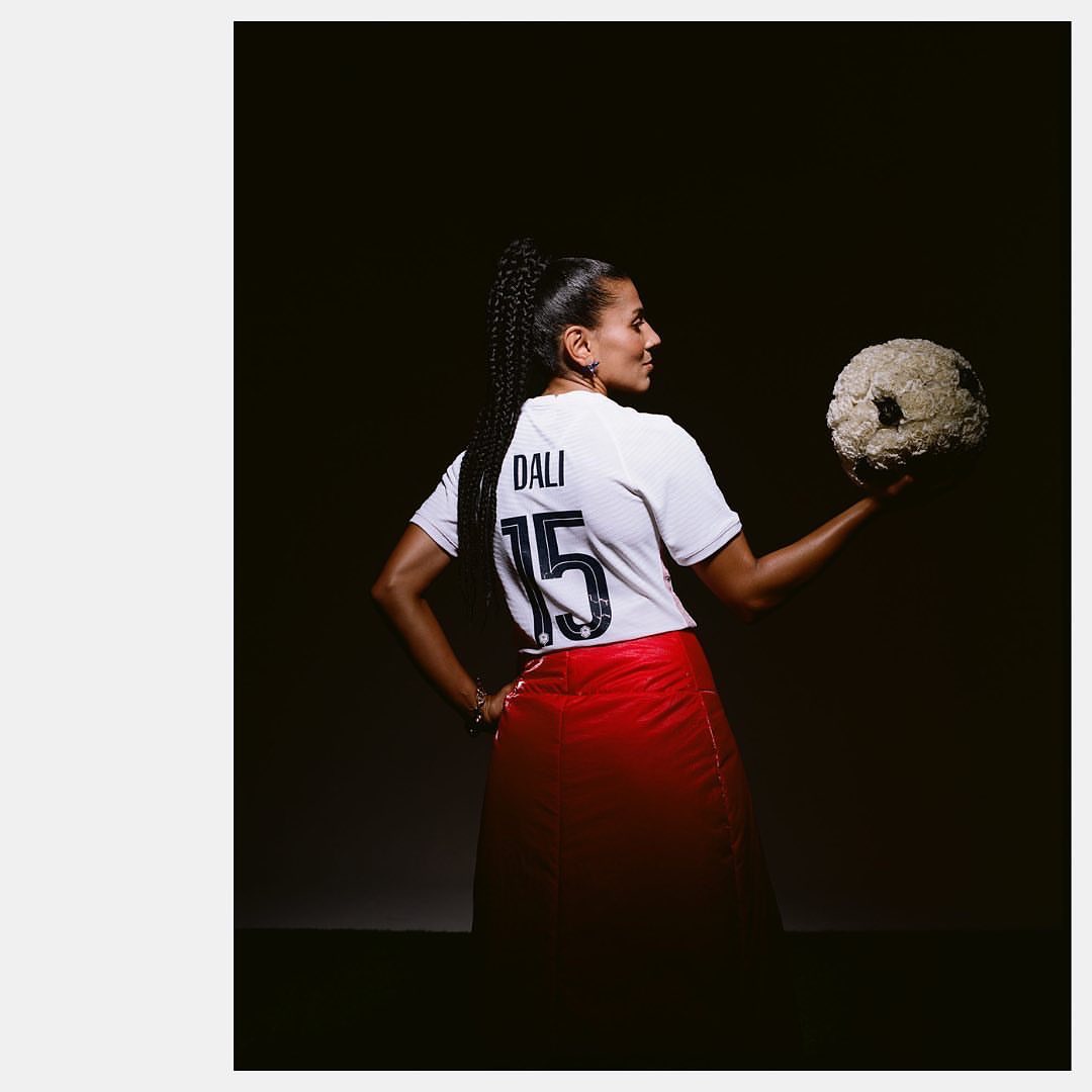 Woman in sports jersey and red skirt holds soccer ball made of flowers in a dark photo studio with overhead lighting, looking to the side.