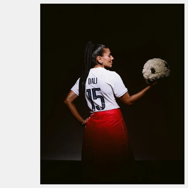 Woman in sports jersey and red skirt holds soccer ball made of flowers in a dark photo studio with overhead lighting, looking to the side.