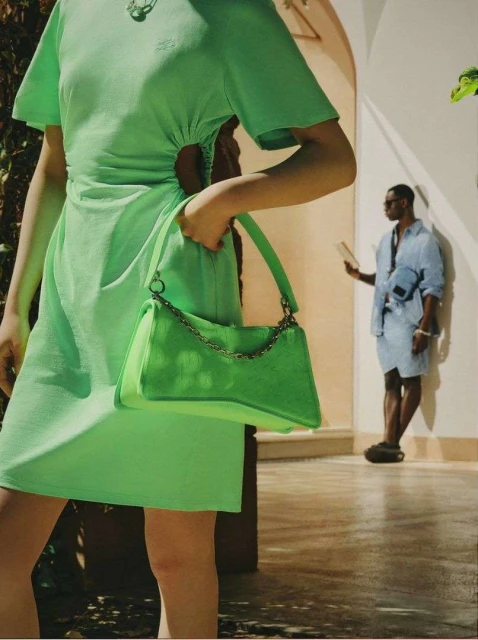 Woman in a lime green dress holds a matching handbag, with a man in blue visible in the background against a light-colored wall.