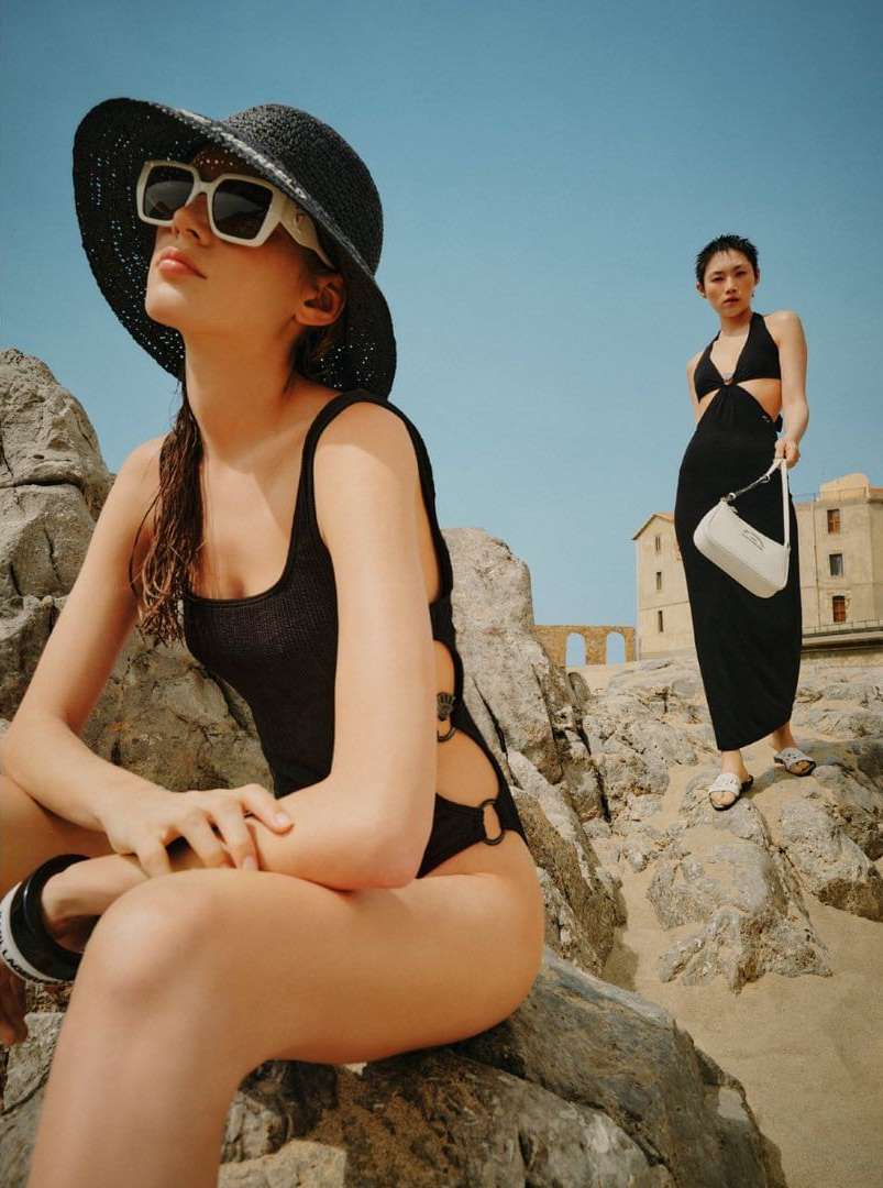 Two women model black swimwear and a cover-up on a rocky beach under a sunny blue sky, showcasing summer fashion and beachwear.