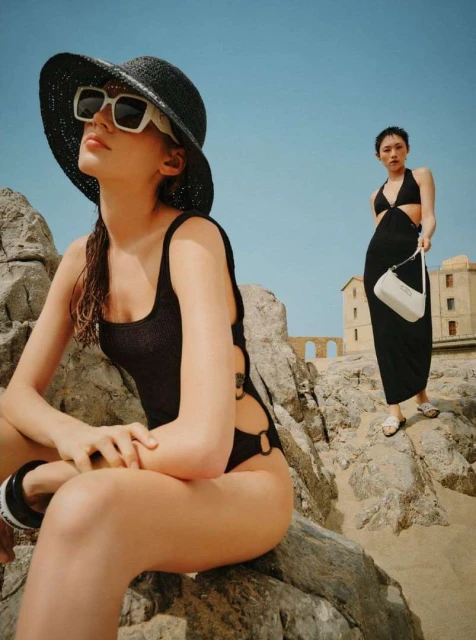 Two women model black swimwear and a cover-up on a rocky beach under a sunny blue sky, showcasing summer fashion and beachwear.