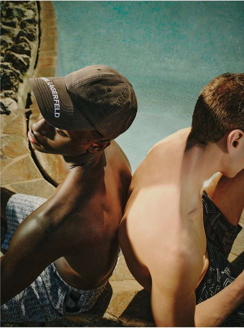 Two shirtless men relax back-to-back near a pool; one wears a Karl Lagerfeld cap. Warm daylight creates highlights and shadows.