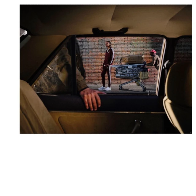 Two men stand outside a car window; one in a tracksuit poses while the other pushes a shopping cart down a brick street.