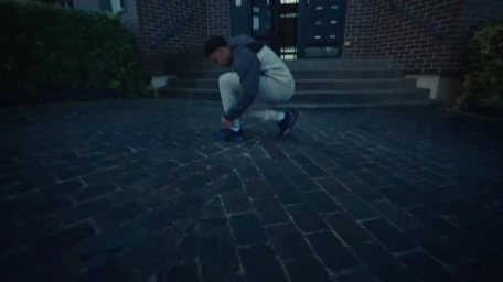 Young man in a tracksuit crouching on a cobblestone courtyard tying his sneakers outside a brick residential building.