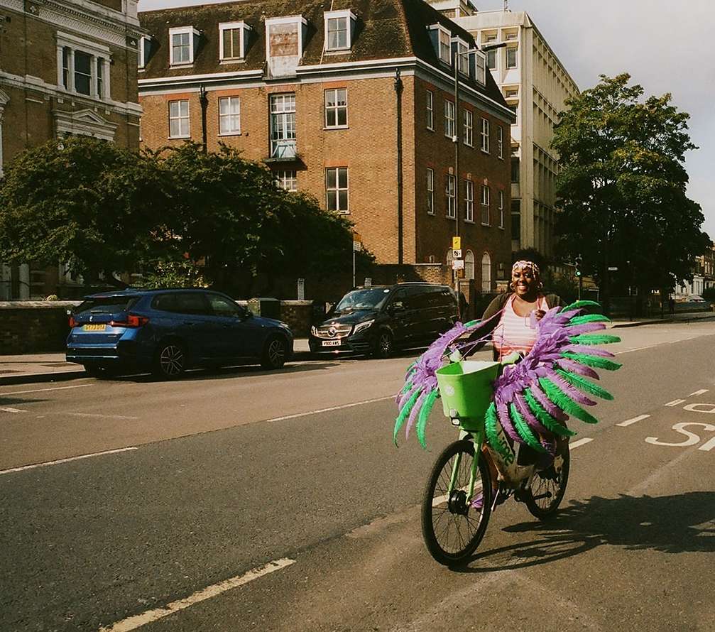 A woman in feathered wings rides a bicycle on a city street near parked cars and buildings in daytime, featuring bright green accents.
