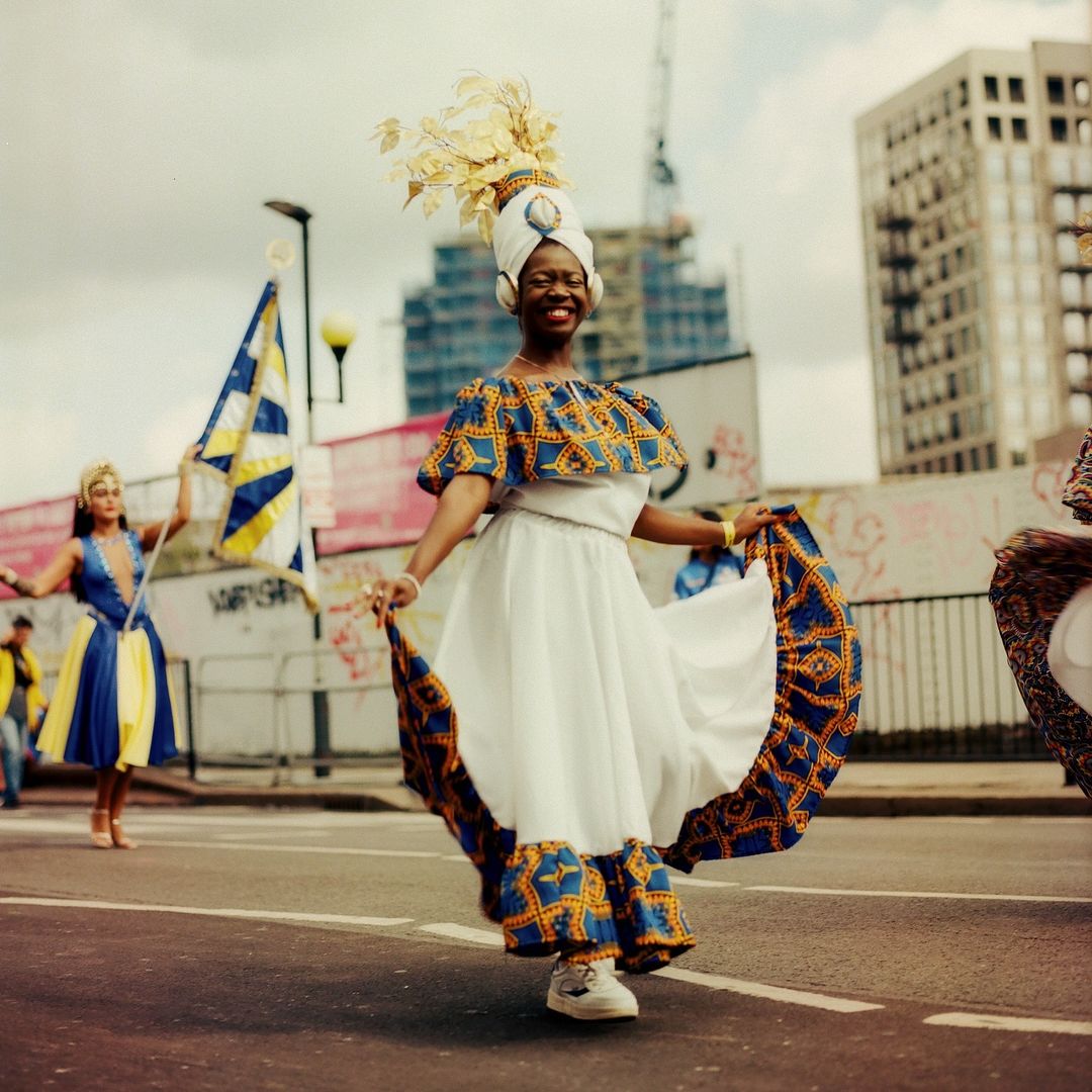 Smiling woman in traditional dress dances in the street during a daytime celebration, with city buildings visible in the background.