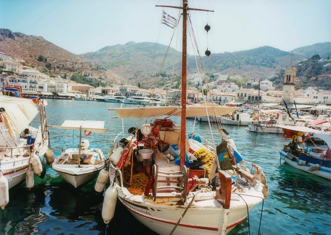 White fishing boats docked in a harbor with clear blue water, nestled by a hillside village under a bright blue sky on a sunny day.