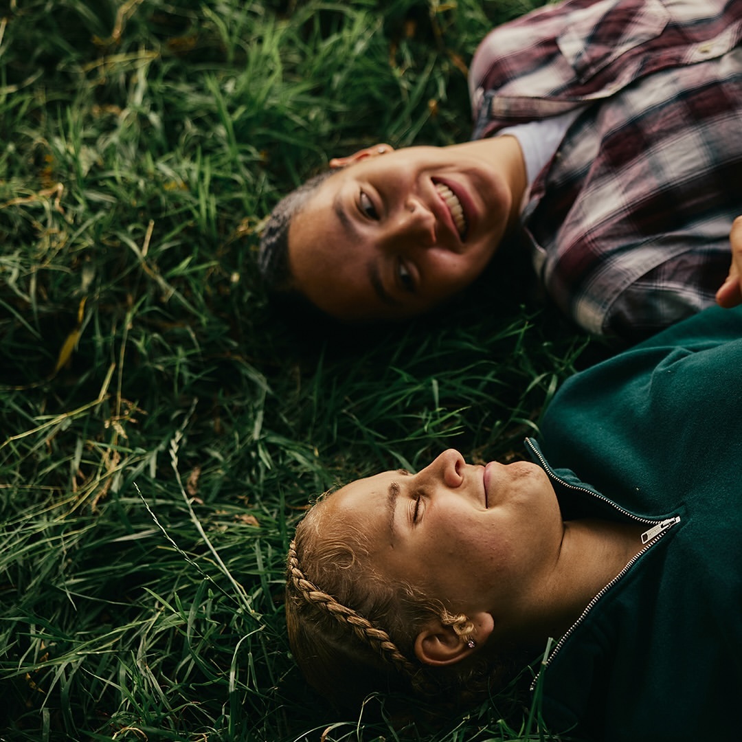 Two women lie side-by-side in green grass, one smiling at the camera while wearing a flannel shirt, the other in a green jacket.