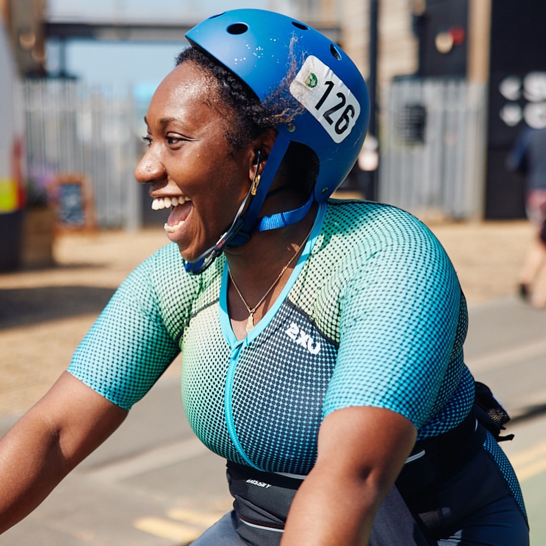Smiling woman wearing a blue helmet and cycling gear rides a bike on a paved road with buildings in the background on a sunny day.