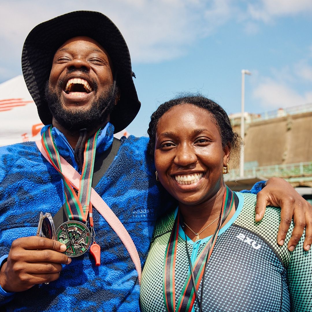 Two athletes, an adult male and an adult female, are posing and smiling outdoors, wearing medals. They appear to be celebrating a victory.