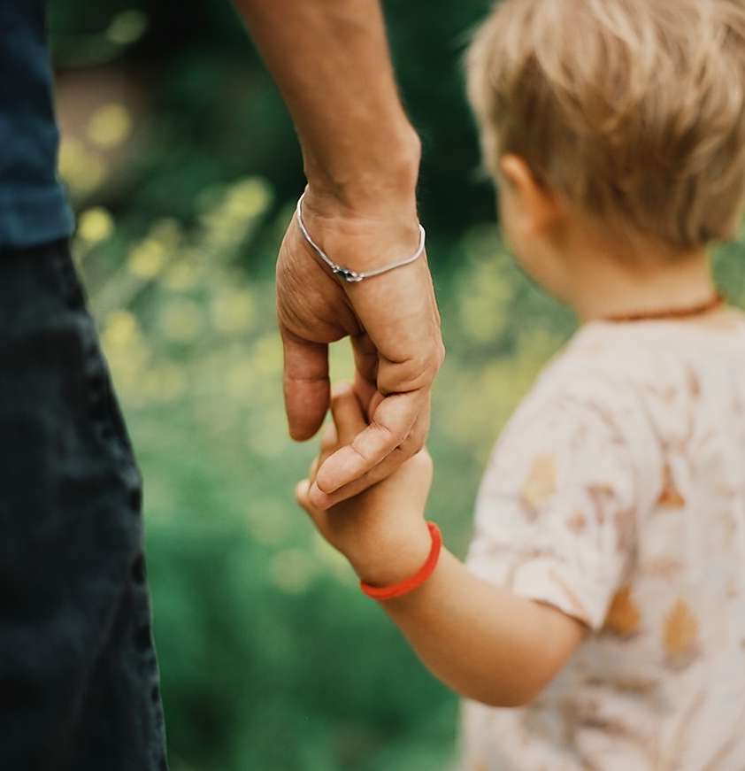 Close-up shot of adult holding hands with a toddler, outdoors in natural green environment. The light is soft and diffused.