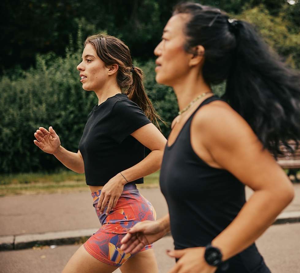 Two women in athletic wear are jogging outdoors on a paved path surrounded by greenery. They are running in unison in daylight.