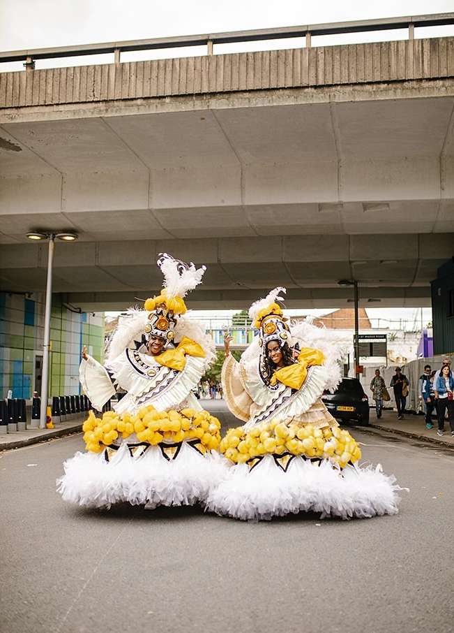 Two people in elaborate yellow and white feathered costumes stand on an asphalt road under a concrete overpass, performing outdoors.