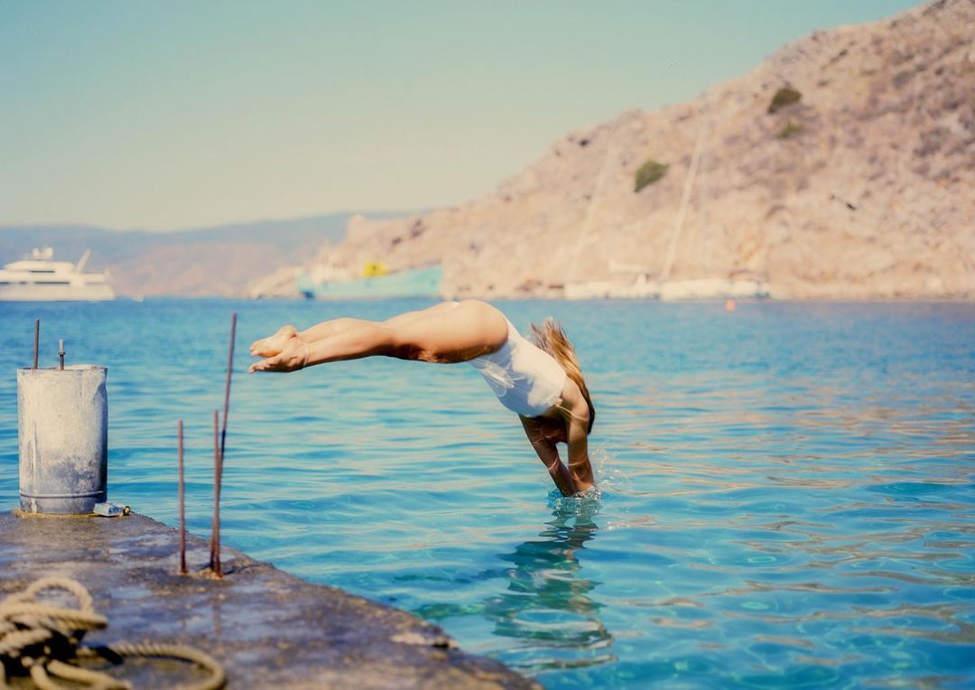 A woman dives into the clear blue ocean from a pier on a sunny day; a boat and mountainous landscape can be seen in the distance.
