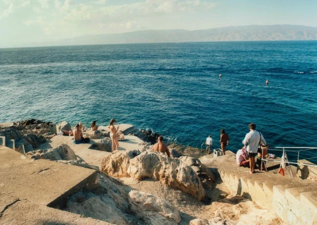 People gathered on a rocky coastal ledge and concrete steps overlooking a wide blue sea under a pale sky, captured by Francis Augusto.