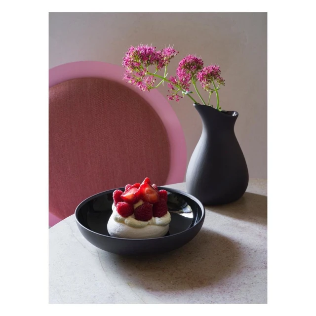 Pavlova dessert with fresh berries, black vase with pink flowers, and pink chair in the background.