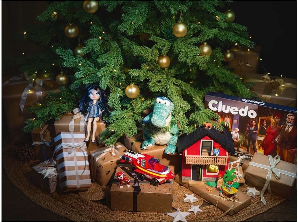 Festive Christmas tree scene with gift-wrapped presents, holiday ornaments, doll, and Cluedo game box underneath, creating a cheerful mood.