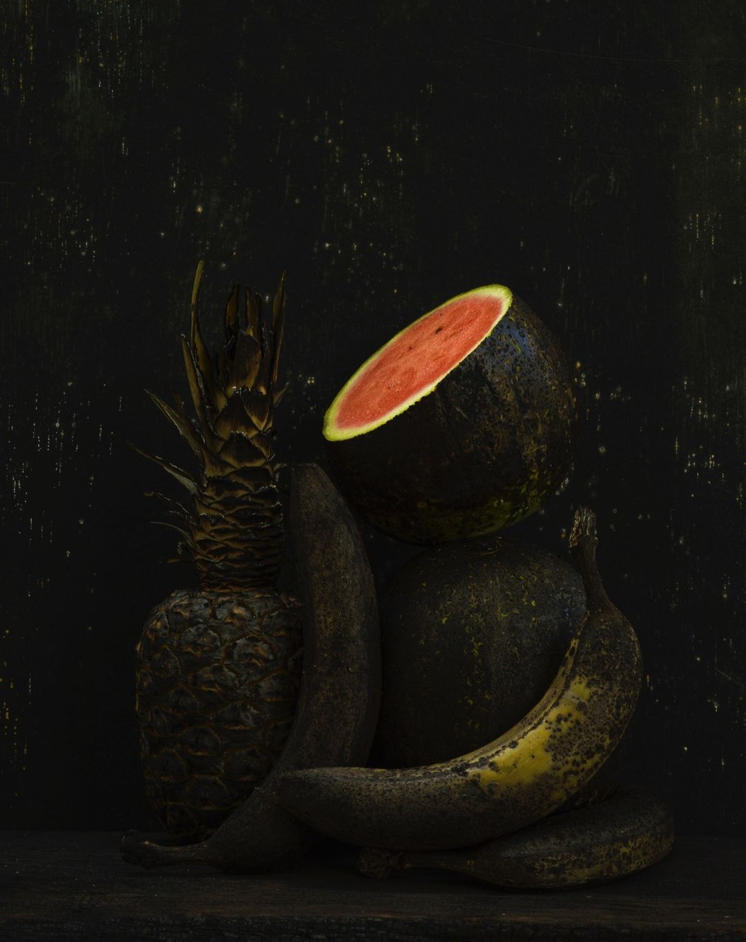 Still life photograph featuring a cut watermelon, whole pineapple, and two bananas arranged against a dark background with textured lighting.