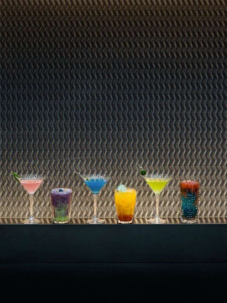 Colorful cocktails lined up on a bar against a textured grey wall, featuring a variety of vibrant drinks in stemmed glasses.