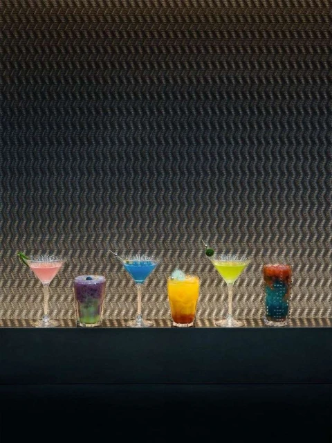 Colorful cocktails lined up on a bar against a textured grey wall, featuring a variety of vibrant drinks in stemmed glasses.