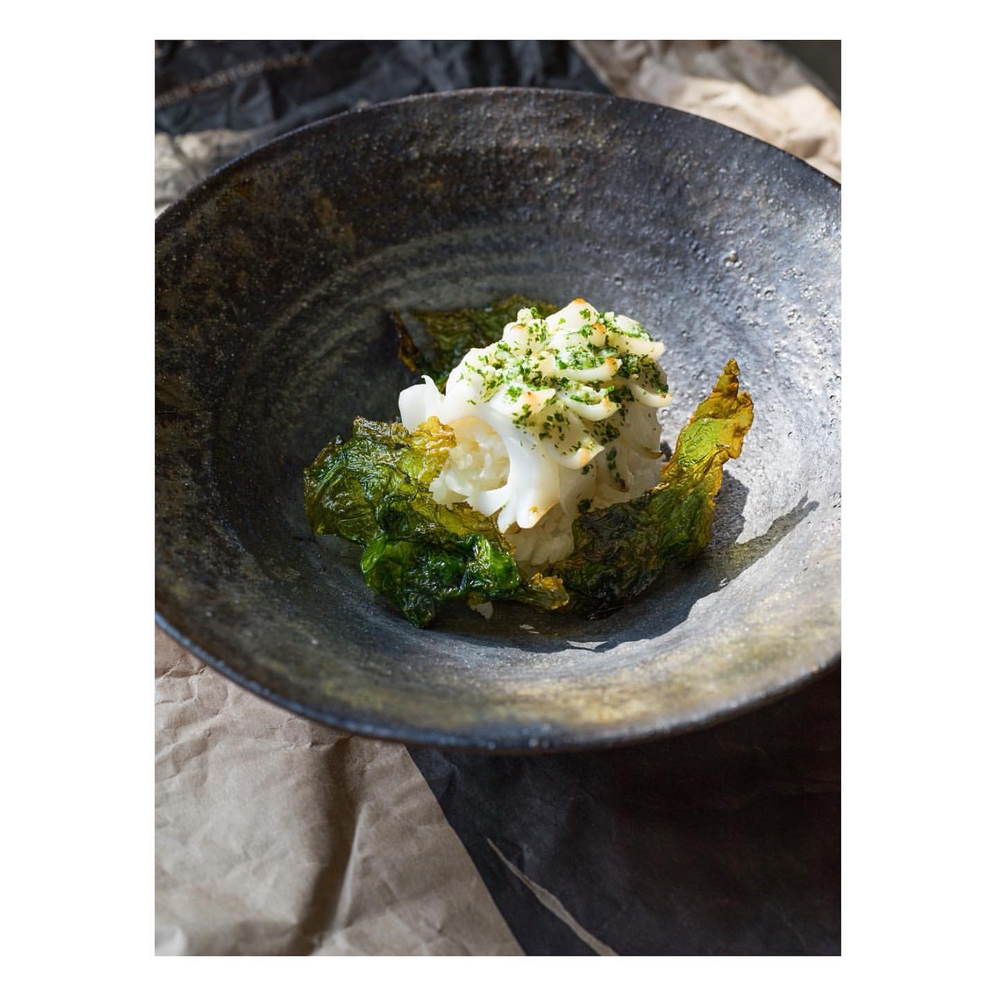 Overhead shot of a black bowl containing an appetizer, with shredded white seafood, green leaves, and a bed of paper in the background.