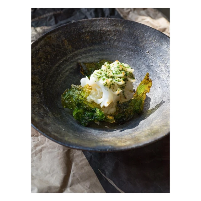 Overhead shot of a black bowl containing an appetizer, with shredded white seafood, green leaves, and a bed of paper in the background.