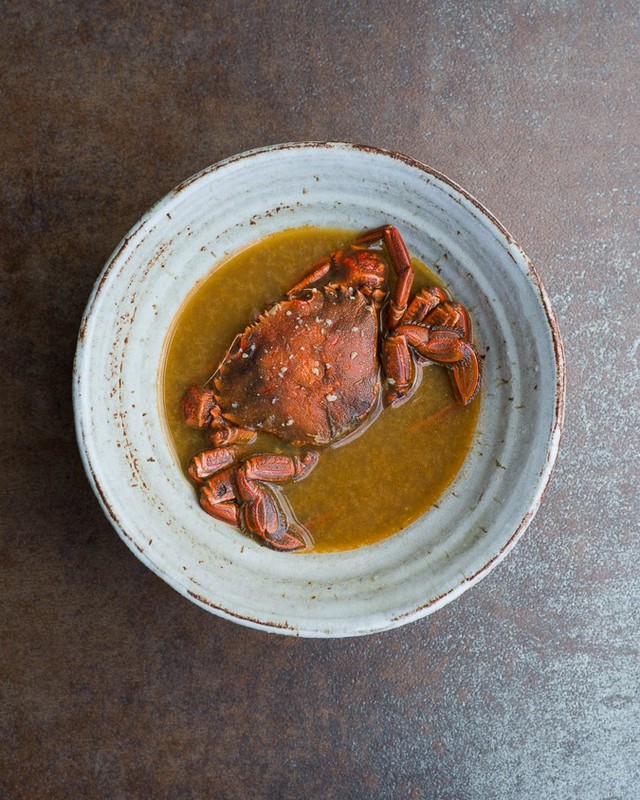Overhead shot of a cooked crab in a light-colored bowl with broth against a mottled brown surface, showcasing food photography.