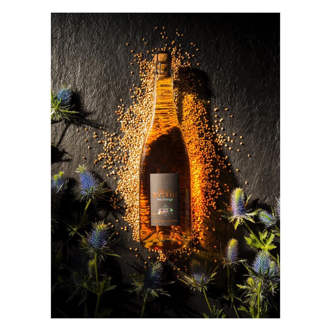 Bottle of light brown liquid with floral decorations and mustard seeds on a dark stone background. Beverage product photography.