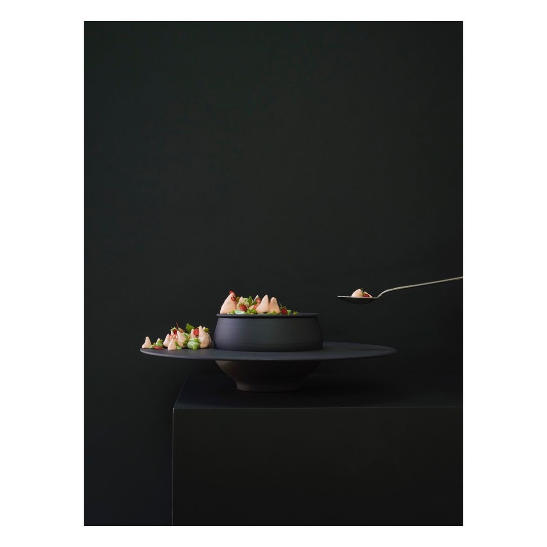 Black matte bowl of salad on a tiered stand being served with a metallic serving spoon against a dark moody monochromatic background.
