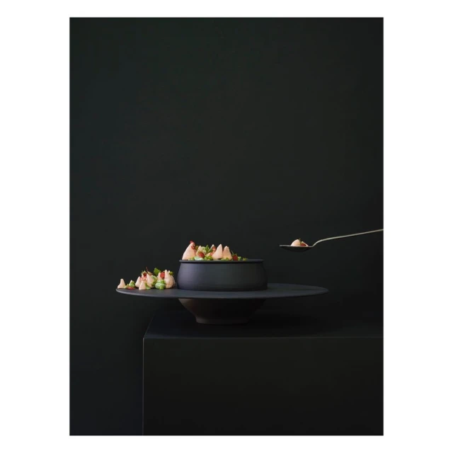 Black matte bowl of salad on a tiered stand being served with a metallic serving spoon against a dark moody monochromatic background.