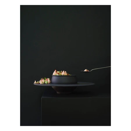 Black matte bowl of salad on a tiered stand being served with a metallic serving spoon against a dark moody monochromatic background.