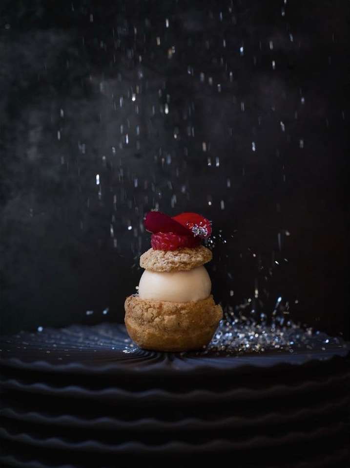 Close-up of a plated dessert featuring a cream puff base, light filling, fresh red berries, and a dusting of glitter against a dark backdrop.