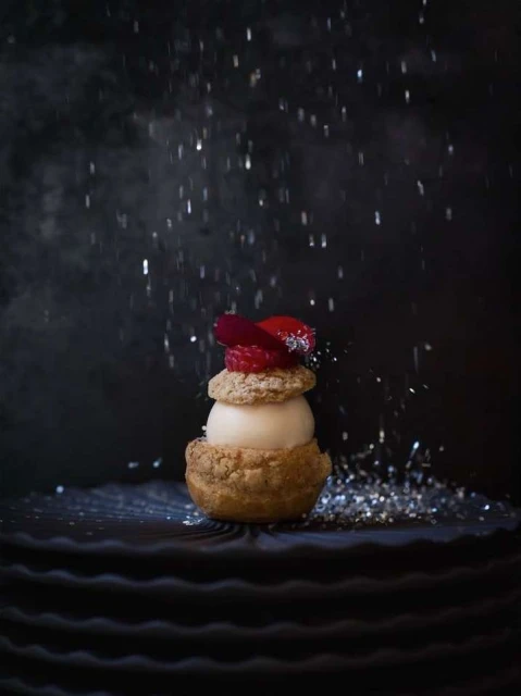 Close-up of a plated dessert featuring a cream puff base, light filling, fresh red berries, and a dusting of glitter against a dark backdrop.