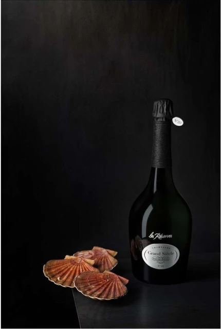 Elegant still life featuring a bottle of champagne with a black foil top, paired with four scallop shells on a dark surface.