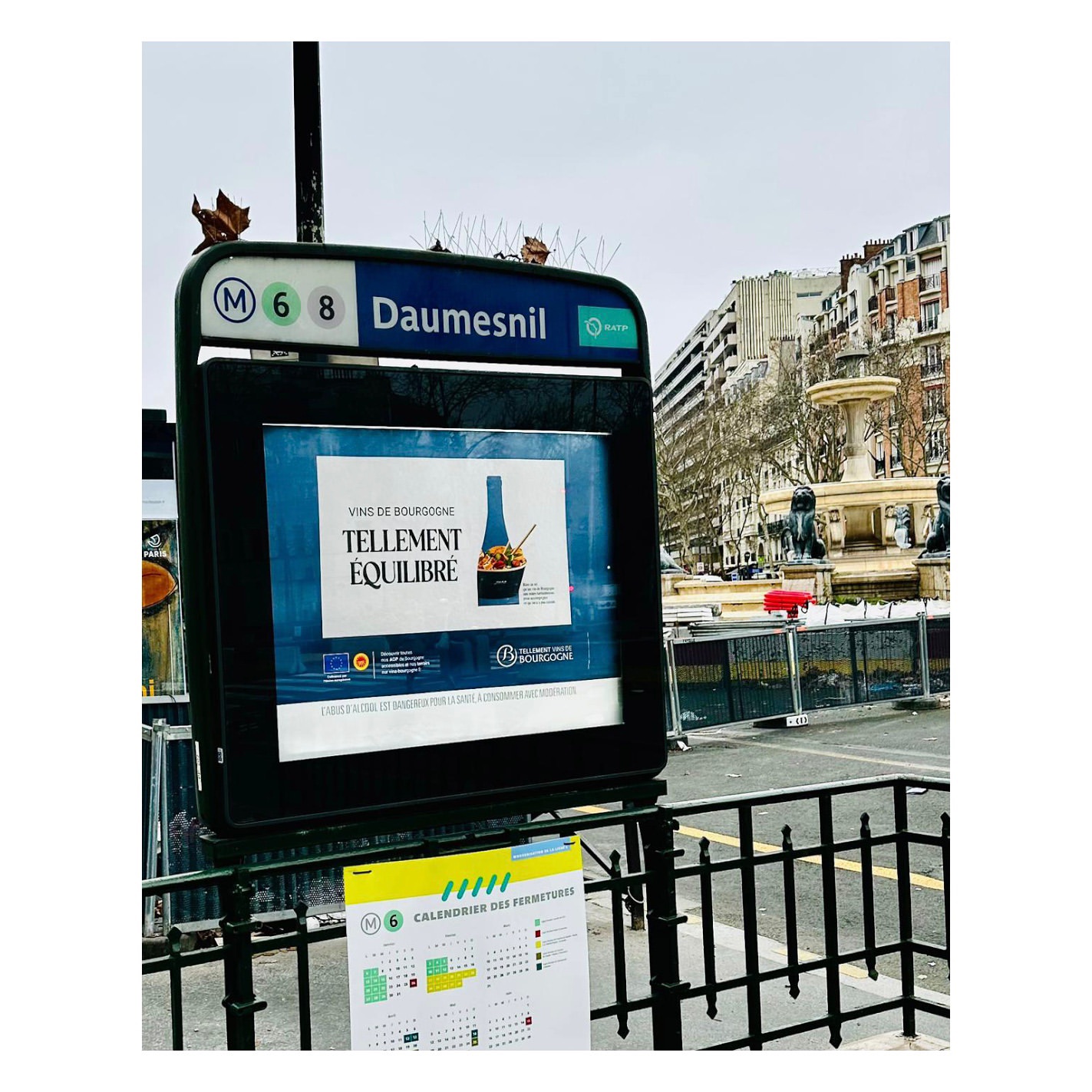 A Paris metro station entrance with an advertisement for wine, featuring a bottle and salad, and a fountain visible in the background.