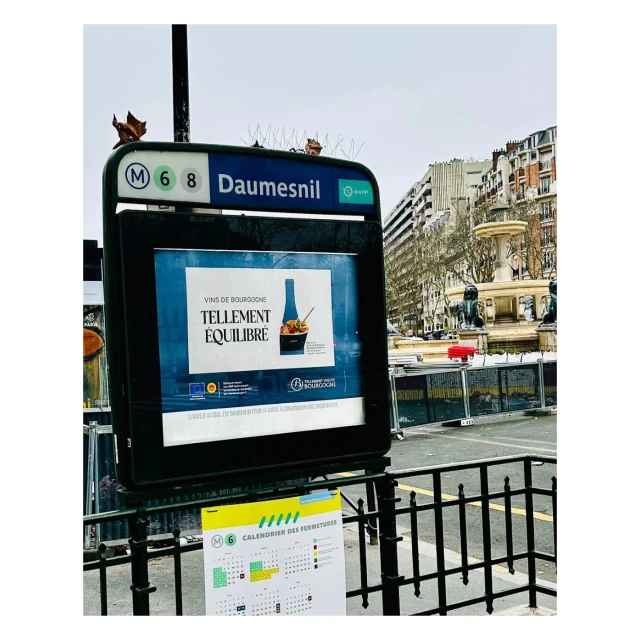 A Paris metro station entrance with an advertisement for wine, featuring a bottle and salad, and a fountain visible in the background.