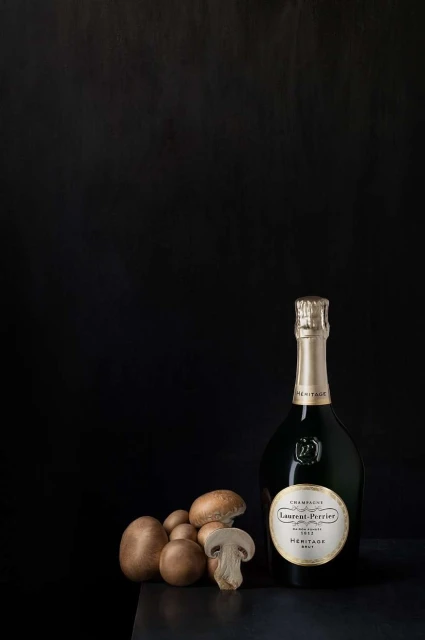 Elegant product shot of a bottle of Laurent-Perrier champagne with a cluster of brown mushrooms on a dark table against a dark background.