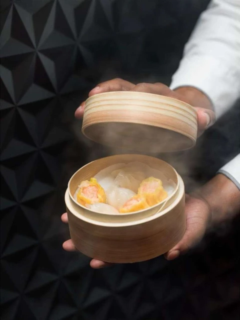 Dim sum in a bamboo steamer held by a person against a geometric black background, showcasing food presentation and culinary photography.