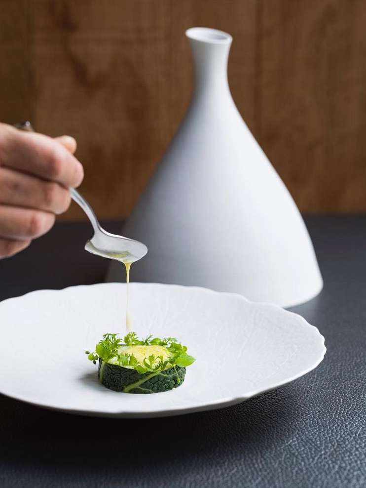 Gourmet food presentation: a single serving of greens on a decorative white plate being drizzled with sauce, set against a neutral backdrop.