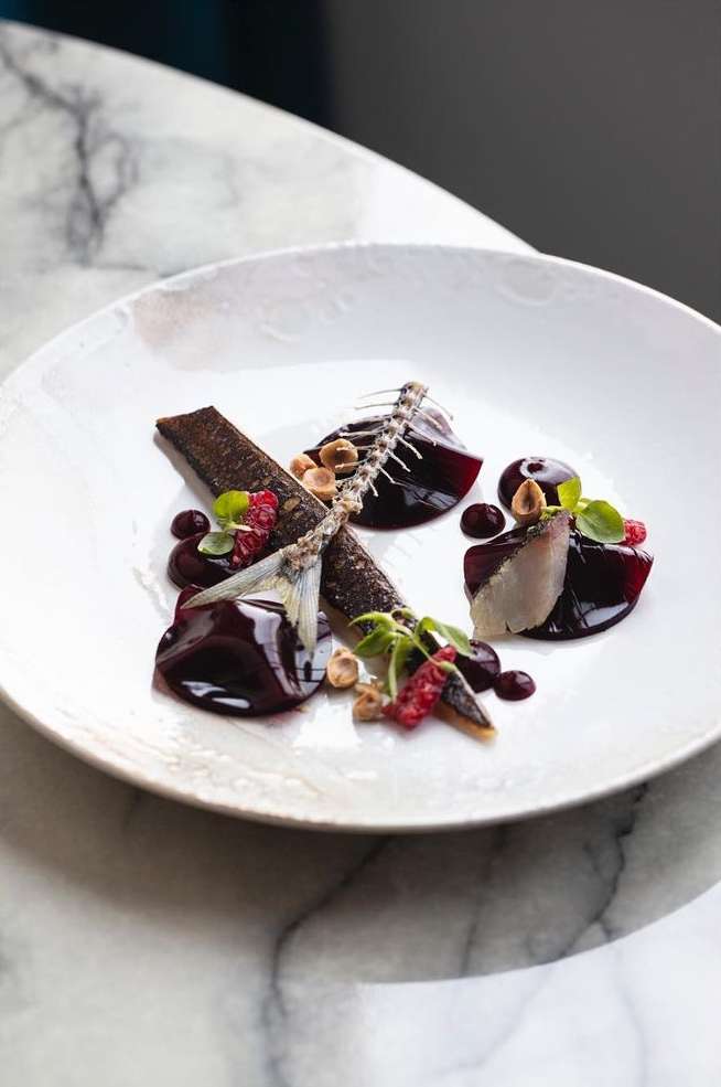 Gourmet food presentation on white plate: creative arrangement of fish, sauce, nuts, and microgreens on a white marble table.