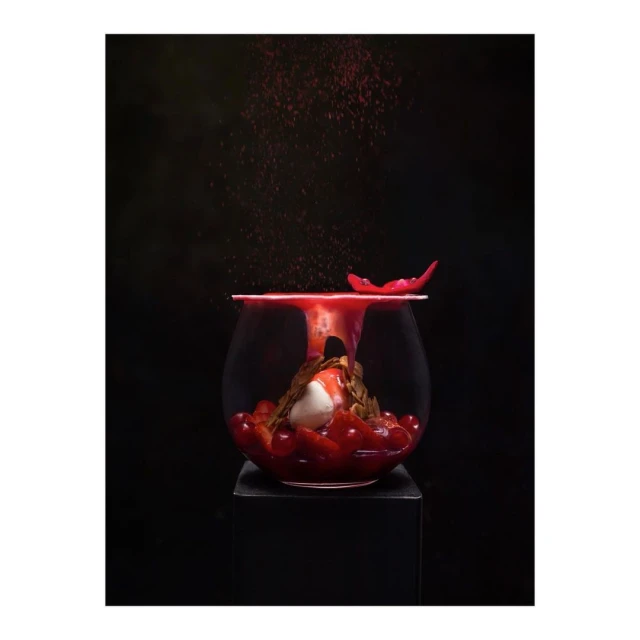 Elevated view of cherry dessert in a glass bowl with rose petals on a black surface against a dark background, dusted with red powder.